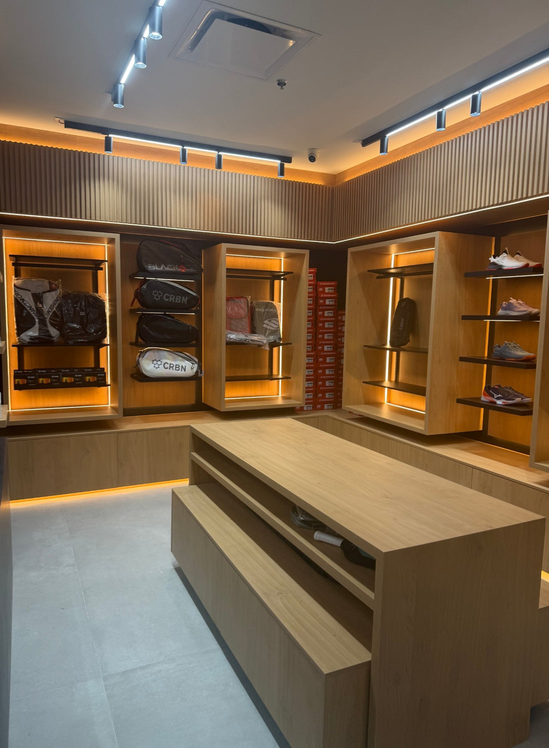 Pickleball bags and court shoes on lit wood shelving at Spinwave Boca, with a long wood display table in front