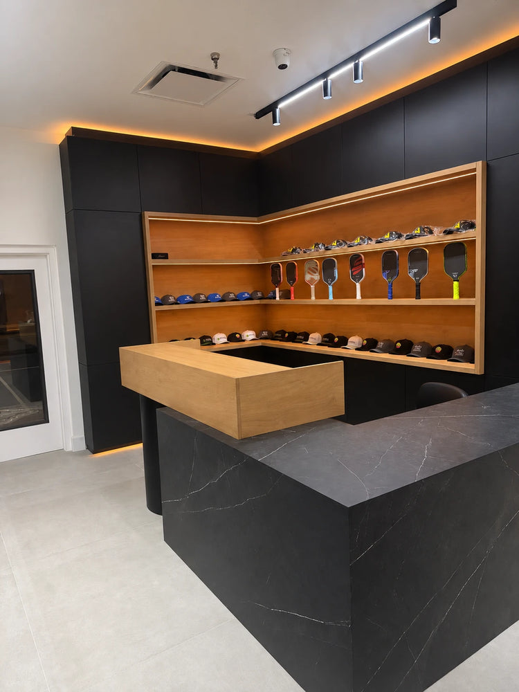 The front counter at Spinwave Boca, with paddles and hats on backlit wood shelves behind a black stone counter