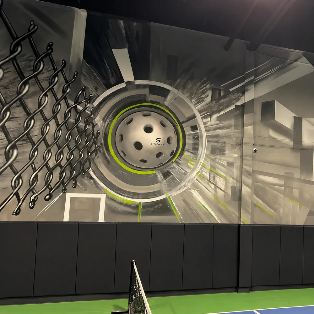 Wall art in The Cage at Spinwave Boca: a giant painted pickleball bursting through a chain-link fence above the padded court wall